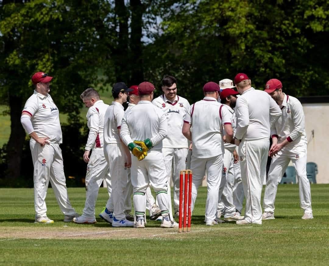 Tewkesbury players in maroon caps gather by the stumps after a wicket